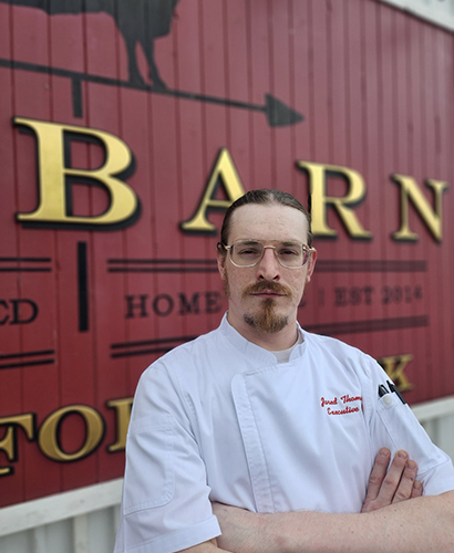 Executive Chef Jared Thompson standing outside next to signage for The Barn restaurant